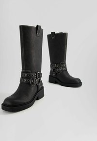 Botas negras hasta la rodilla hechas de material texturizado, con dos correas con hebillas y detalles metálicos, y un tacón grueso con plataforma.