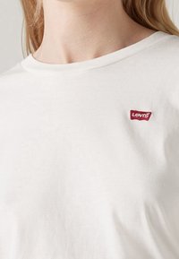 Personne portant un t-shirt blanc Levi's avec un petit patch rouge avec le logo Levi's sur la partie supérieure gauche de la poitrine.