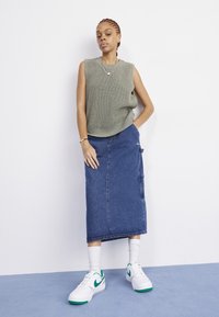 Donna in piedi contro un muro bianco, indossa un top grigio in maglia senza maniche, una lunga gonna di jeans, sneakers bianche con dettagli verdi e calze bianche.
