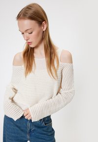 Witte, gebreide trui met een off-shoulder ontwerp, textuurpatroon en lange mouwen. Gecombineerd met hooglopende spijkerbroeken.