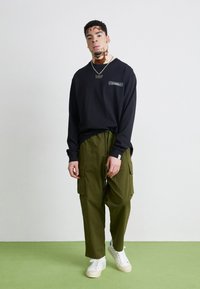 Schwarzes Langarmshirt mit Markenlogo, olivgrüne Cargo-Hose und weiße Sneaker. Weiße Sohle mit strukturierten Akzenten, entspannte Passform der Hose.