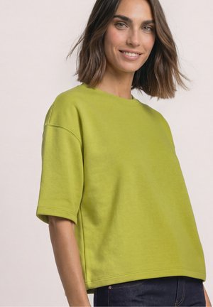 Kvinde med skulderlangt brunt hår, smilende, iført en løs limegrøn kortærmet top og mørkeblå jeans mod en ensfarvet baggrund.