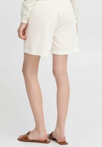 Shorts en lin blancs avec une taille élastique, présentant une coupe décontractée et un ourlet roulé. Associés à des sandales plates marron.
