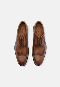 Des chaussures en cuir marron à lacets présentent un design brogue avec des accents perforés et une finition lisse, montées sur une semelle en caoutchouc noir.