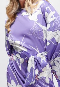 Lila Blumenkleid mit großen weißen Lilien und rosa Akzenten. Verfügt über einen lockeren Schnitt, weite Ärmel und ein gebundenes Taillendetail. Glatte Stoffstruktur.