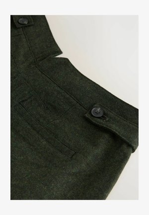 Tessuto verde scuro con un aspetto testurizzato, presenta una chiusura con bottone e una tasca laterale in vita, evidenziando un design su misura.