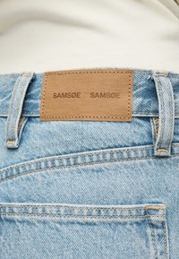 Jean en denim bleu clair avec une étiquette en cuir fauve cousue au-dessus de la poche arrière, affichant le nom de la marque "SAMSOE SAMSOE".