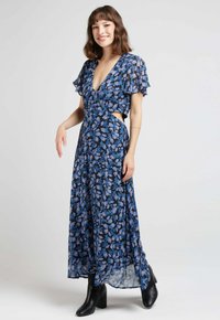 Robe longue à fleurs bleues avec un décolleté en V profond, des manches volantes, des côtés découpés et une jupe fluide. Assortie avec des bottines noires à talons.
