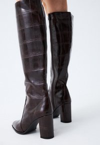 Botas marrones hasta la rodilla, hechas de piel de cocodrilo con estampado brillante, con un tacón ancho y un cierre con cremallera lateral.