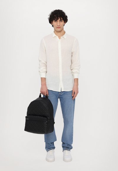 Sac à dos noir matelassé avec un sommet arrondi, en tissu texturé, poche zippée à l'avant et bretelles ajustables, tenu par une personne en tenue décontractée.