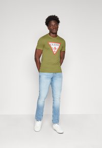 T-shirt verde oliva a maniche corte con un logo triangolare rosso raffigurante un punto interrogativo. Abbinato a jeans blu chiaro e sneakers bianche.
