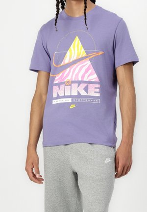 Mann in lila Nike T-Shirt mit gelbem und pinkem dreieckigem Design und grauen Nike-Sweatpants, der vor einem weißen Hintergrund steht.