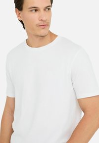 Junger Mann mit kurzen, dunklen Haaren, der ein schlichtes weißes T-Shirt mit kurzen Ärmeln trägt und nach links blickt, vor einem weißen Hintergrund.