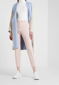 Manteau à couches bleu clair et beige porté sur un col roulé blanc, assorti à un pantalon court rayé rose et des chaussures de sport blanches.