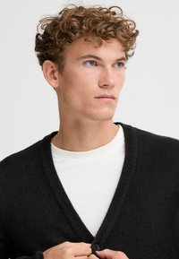 Svart strikket cardigan med dyp V-hals, knappelukking og ribbestrikkede kanter. Brukt over en hvit crewneck skjorte. Myk tekstur synlig.