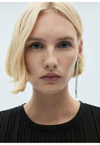 Blond lasje s teksturiranim bobom, oblečen v črn naguban top in dolge srebrne uhane. Nevtralna ličila z vidnimi pegami.