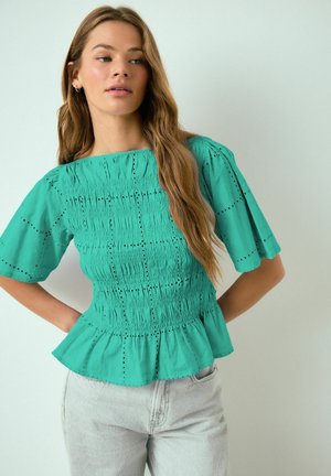 Junge Frau mit langen, welligen Haaren, die eine türkisfarbene, geraffte Bluse mit Lochstickereien und kurzen Ärmeln trägt, kombiniert mit hellblauen Jeans.