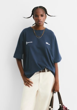 T-shirt oversize bleu marine avec le texte blanc "OSE PLUS" et "CRÉEZ DES SOUVENIRS". Associé à un pantalon clair et un sac à main marron.