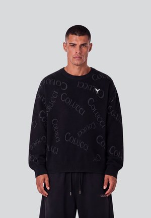 Mann trägt einen schwarzen Pullover mit dem wiederholt aufgedruckten Wort "Colucci" und passende schwarze Hose vor einem schlichten grauen Hintergrund.