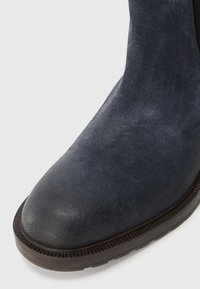 Marinblå mocka ankelnboot med rund tå och texturerad yta, med en brun gummisula med ribbat mönster.