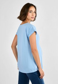 Ljusblå topp med kort ärm och rundad halsringning, mjukt tyg, avslappnad passform och något längre bakstycke, i kombination med mörka jeans.