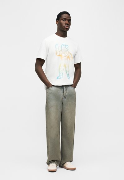 Άνδρας με χέρια στις τσέπες, φορώντας λευκό μπλουζάκι με πολύχρωμο γραφικό αστροναύτη, φαρδιά ξεθωριασμένα γκρίζα τζιν και λευκά αθλητικά παπούτσια.