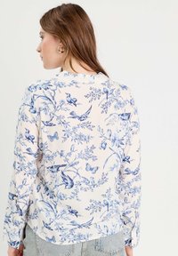 Blusa a maniche lunghe color crema con stampa floreale e di uccelli blu, dotata di scollo con volant e polsini a bottone. Tessuto morbido e leggero.