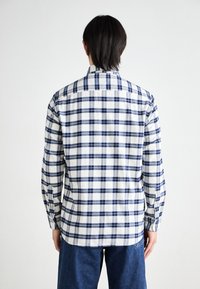 Person mit dunklen Haaren, der ein langärmliges, kariertes Hemd in Weiß und Marineblau sowie Blue Jeans trägt, von hinten auf einem weißen Hintergrund betrachtet.