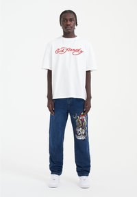 Camiseta gráfica blanca con texto en rojo "E.D. Hardy", combinada con jeans de mezclilla azules que presentan un diseño colorido de calavera y serpiente bordado.