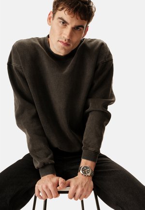 Unge mand i sort sweatshirt og jeans sidder på en skammel, hænderne griber fat i sædet, iført et sølvur med sort urskive.