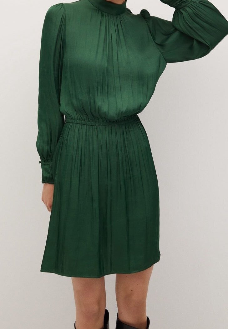 Femme portant une robe plissée vert foncé à manches longues, avec un col montant et une taille cintrée, associée à des bottes noires.