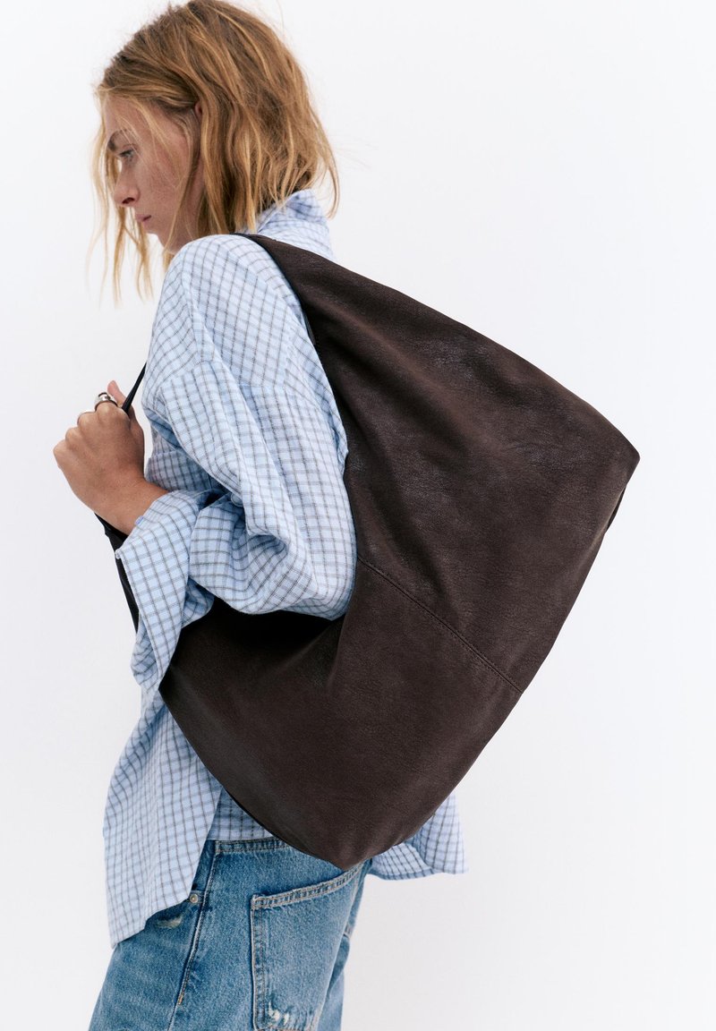 Femme portant une chemise bleue à carreaux et un jean, portant un grand sac à bandoulière en cuir marron foncé sur fond blanc.