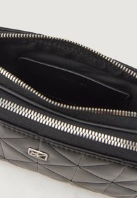 Prošívaná černá kožená taška se stříbrným zipem. Uvnitř se nachází hladká černá podšívka a viditelná sekce na karty.