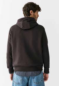 Brązowa bluza z kapturem, wykonana z miękkiego materiału, z regulowanym kapturem, ściągaczami na rękawach i u dołu. Połączona z koszulą w paski i jasnoniebieskimi dżinsami.