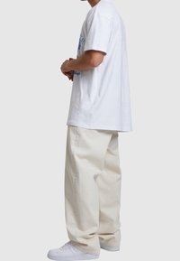 Bílé oversized tričko s modrou grafickou potiskem, v kombinaci s krémovými volnými kalhotami a bílými teniskami.