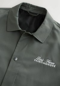 Veste vert foncé avec boutons-pression et col, avec texte brodé blanc "Paris - France SAINT-HONORÉ" sur la poitrine.