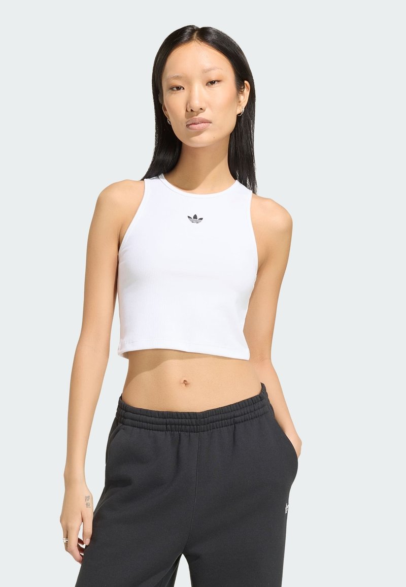 Aziatische vrouw met lang zwart haar, draagend een wit cropped tanktop en zwarte sweatpants, staand met één hand in de zak.