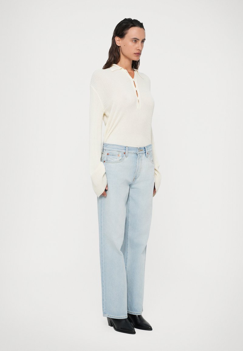 Pull en maille côtelée blanc cassé avec col, patte de boutonnage, associé à un jean légèrement bleu avec une coupe ample et des bottines noires à bouts pointus.