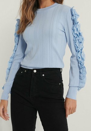 Woman wearing a fitted light blue sweater with vertical knit patterns and ruffled sleeves paired with high-waisted black jeans.