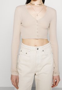 Top beige à manches longues raccourci en côte avec un décolleté en V, doté d'un rang de six boutons ronds sur le devant. Associé à un jean blanc taille haute.