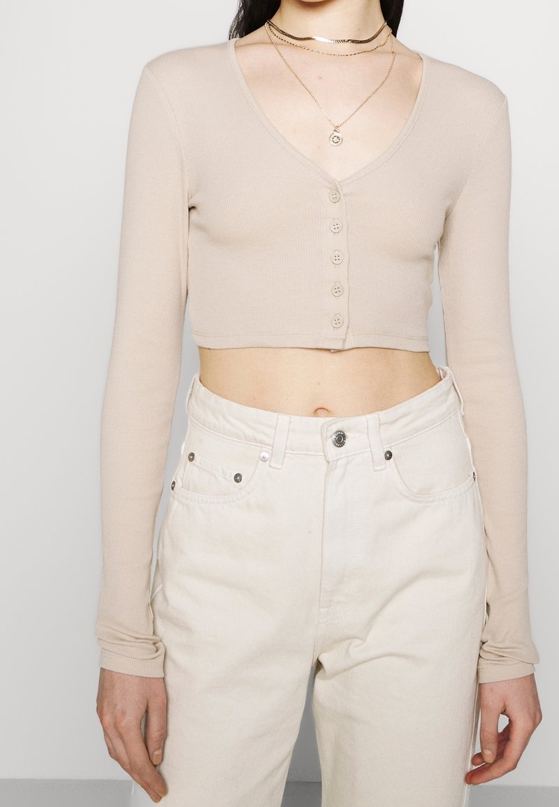 Top beige à manches longues raccourci en côte avec un décolleté en V, doté d'un rang de six boutons ronds sur le devant. Associé à un jean blanc taille haute.