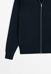 Cardigan zip-up blu navy realizzato in tessuto morbido. Presenta maniche lunghe, polsini a coste e una cerniera argentata che scorre al centro.