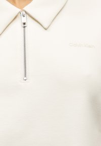 Κοντινό πλάνο λευκού-μονόχρωμου μακρυμάνικου τοπ με γιακά, ασημένιο φερμουάρ και διακριτικό λογότυπο Calvin Klein στο στήθος.