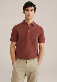 Kortærmet, ribbet polo-skjorte i rustfarve med tre-knaps placket. Stoffet har en struktureret overflade. Bæres med lys beige bukser.