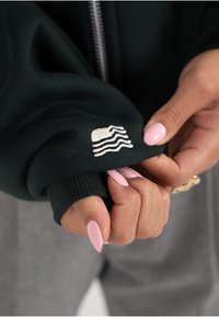 Mørkegrønn hettegenserstoff med ribbede mansjetter, med en hvit brodert logo av bølger. Nærbilde som viser hender som holder ermet.