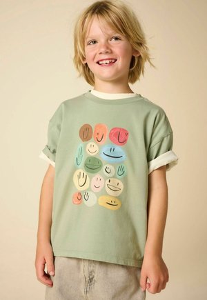 Kind met blond haar glimlachend, draagt een lichtgroen T-shirt met kleurrijke abstracte smileygezichten en beige broek, staand tegen een effen achtergrond.