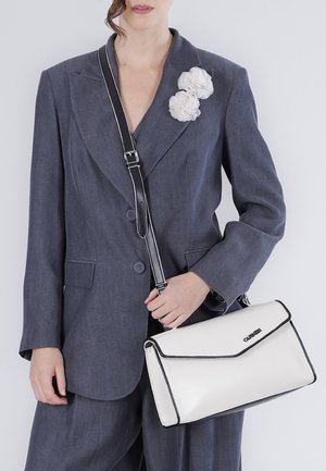 Femme en tailleur gris avec deux fleurs en tissu blanc sur le revers, tenant un sac bandoulière rectangulaire blanc avec des bordures noires et une sangle ajustable.