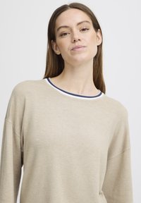 Jeune femme aux longs cheveux bruns portant un pull beige avec un col rayé blanc et marine, se tenant devant un fond blanc uni.