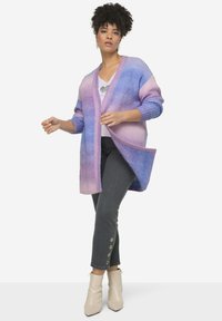 Överdimensionerad cardigan med en övergång från lavendel till blått, ribbad textur, öppen front och långa ärmar, kombinerad med grå byxor och beige stövlar.