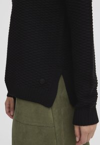 Czarny dziergany sweter z fakturowanym wzorem, ściągaczami na mankietach i bocznymi rozcięciami. Zawiera okrągły czarny guzik przy krawędzi.